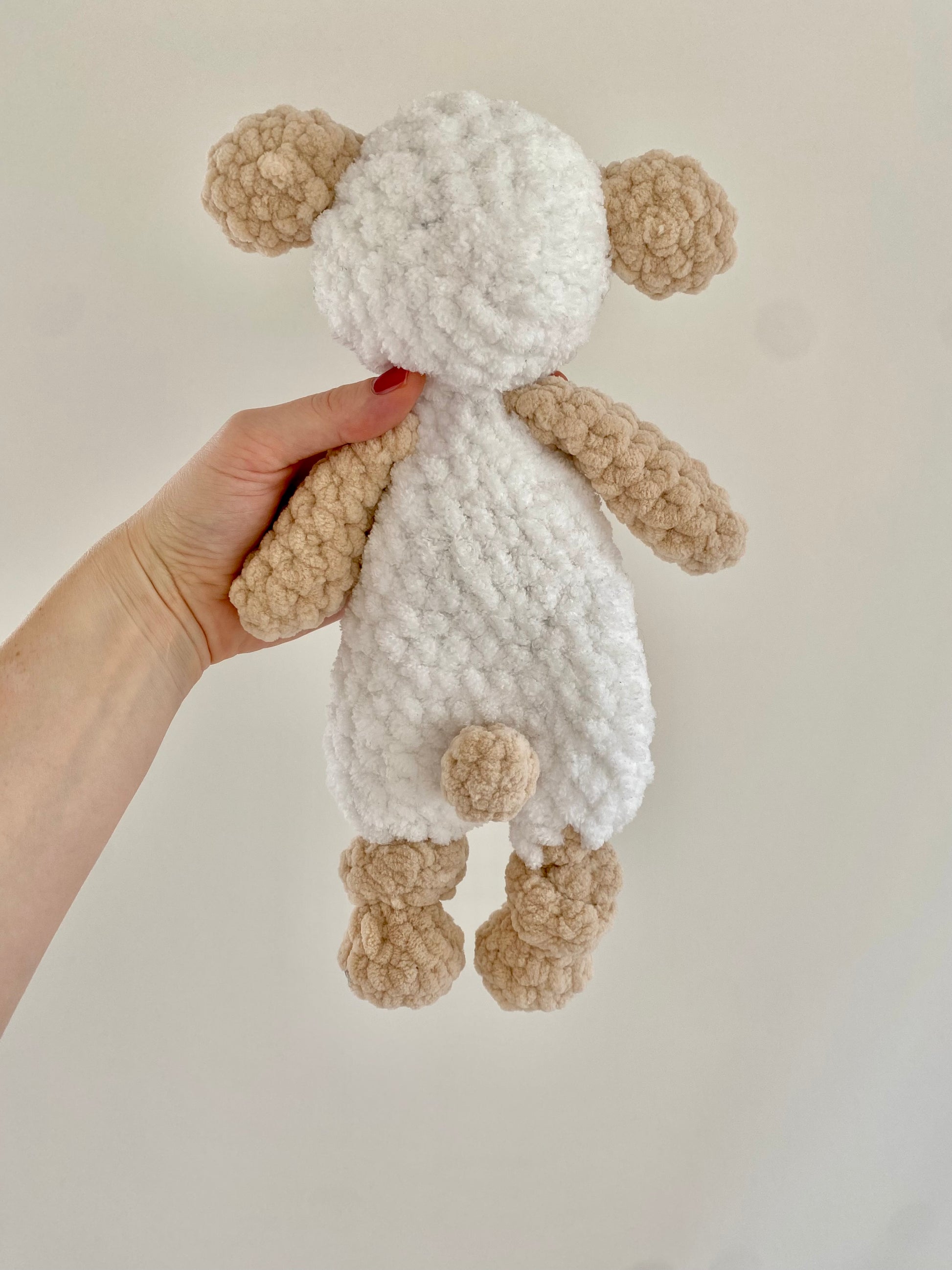 The back view highlights the carefully crafted fluffy texture of Lou the Lamb’s body, created with soft yarn to replicate a sheep’s wool, along with the knotted lovey design for added comfort and charm.